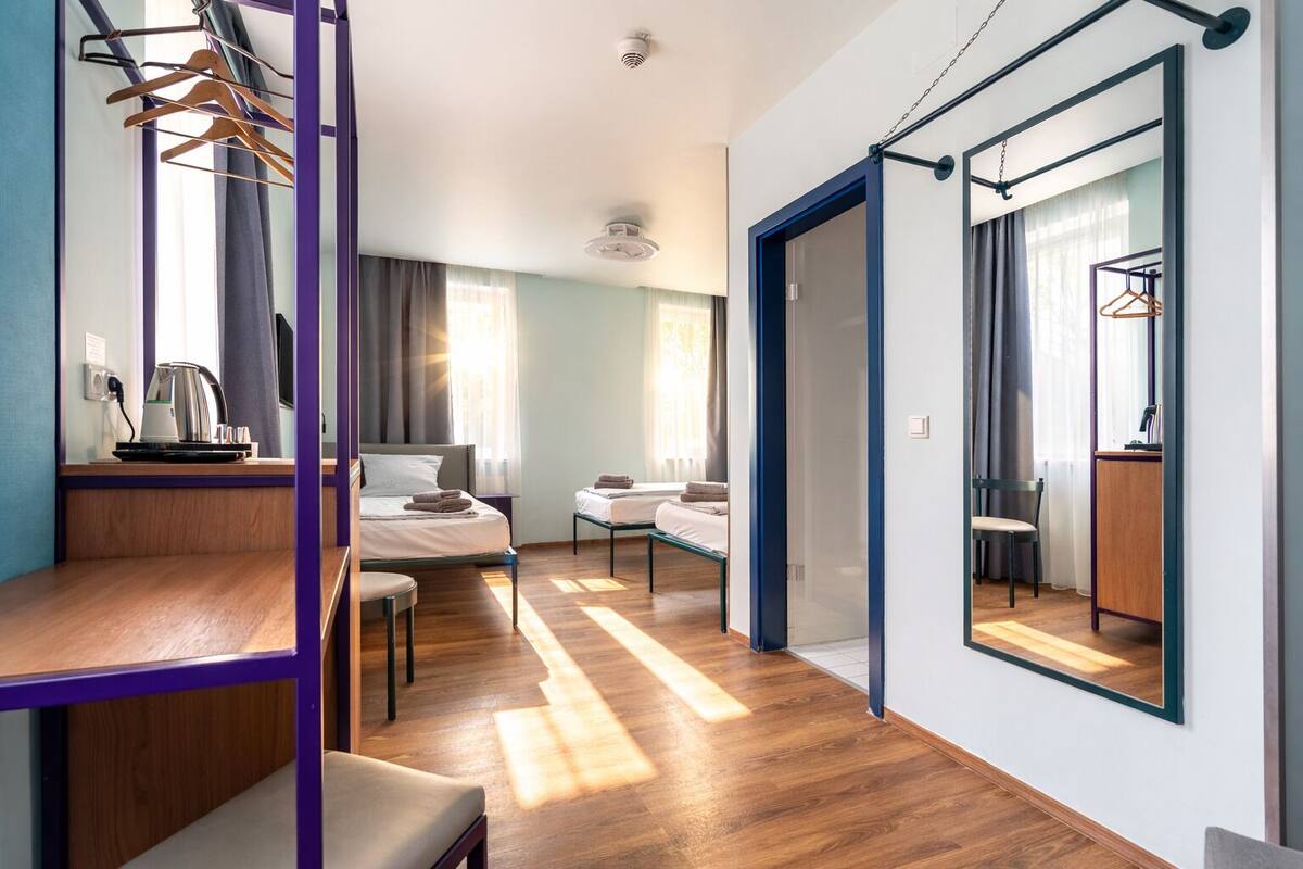 A modern hotel room with a wooden desk, two beds, a wardrobe, and a large mirror.