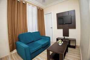 Un salon avec un canapé de couleur turquoise, une table basse en bois surmontée d’un vase, un téléviseur à écran plat fixé au mur et une porte à cadre blanc.