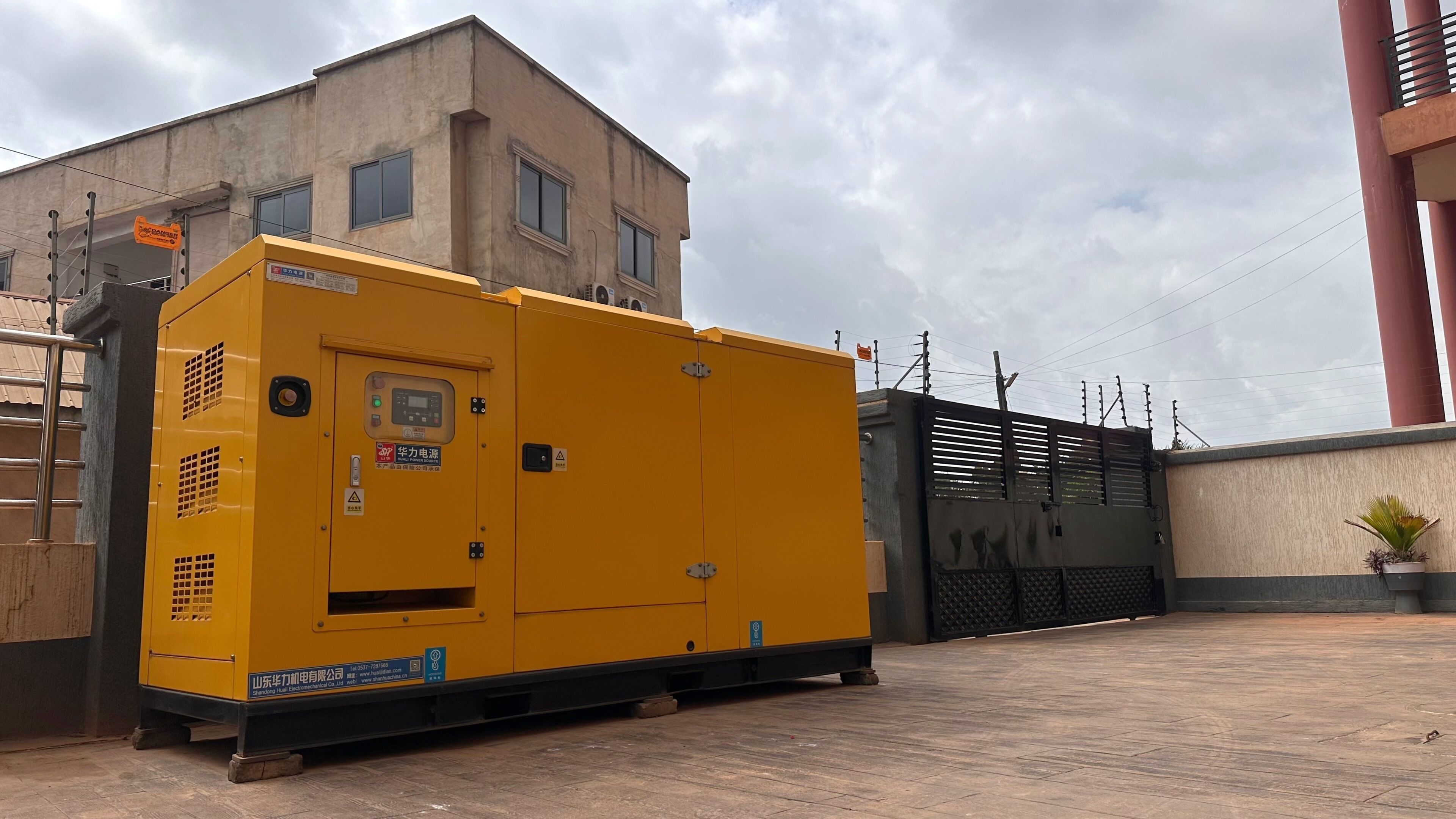 A large yellow industrial generator with a control panel, situated outdoors next to a building with a gated entrance.