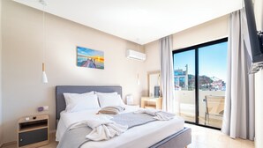 A modern hotel room with a large bed, a nightstand, a bedside table, a window with a view of buildings, and a balcony. - Blue Wave Suites - Ixia (Rhodes)