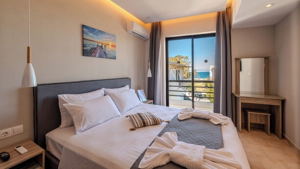 A hotel room with a bed, a nightstand, a desk, a large window with a view, and an air conditioning unit. - Blue Wave Suites - Ixia (Rhodes)
