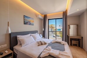 A hotel room with a bed, a nightstand, a desk, a large window with a view, and an air conditioning unit. - Blue Wave Suites - Ixia (Rhodes)