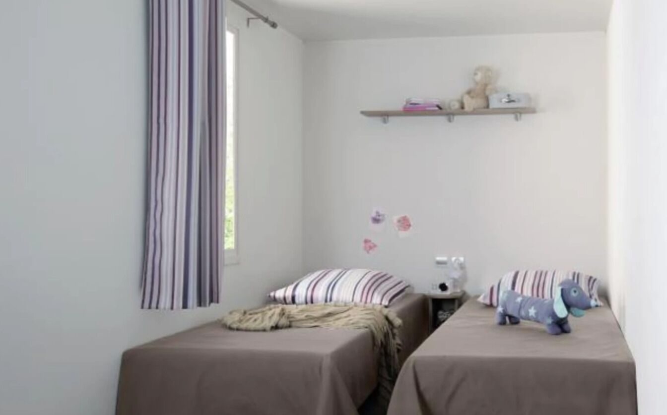 Un dormitorio con dos camas, cortinas a rayas, una estantería con objetos decorativos y un peluche sobre una de las camas.