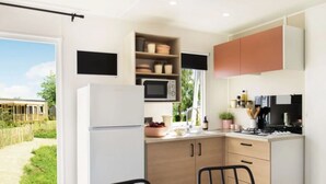 A modern kitchen with a refrigerator, microwave, and stove. There is a dining table with chairs and a window overlooking a garden. - Résidence Santa (Ghisonaccia)