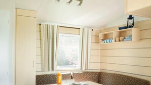 A compact kitchen with a dining area, featuring a window with curtains, a small shelf with items, and a bench with a polka dot cushion. - Résidence Santa (Ghisonaccia)