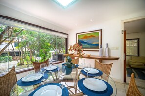 Uma sala de jantar com mesa de vidro posta para seis pessoas, rodeada por cadeiras de vime. Há um quadro na parede acima da mesa.