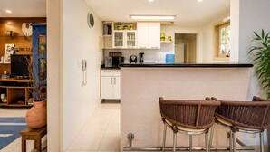 A compact kitchen with a tiled backsplash, a coffee machine, and a water dispenser. There are two wicker bar stools in front of the counter.