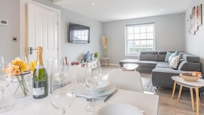 A modern living room with a grey sofa, a coffee table, and a dining area set with wine glasses and a bottle. - 4-Bedroom Seafront House in Southend | Sea Views - Pass the Keys (Southend-on-Sea)