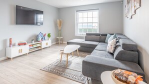 A modern living room with a grey sectional sofa, a flat-screen TV, a small white table, and a window with a view. - 4-Bedroom Seafront House in Southend | Sea Views - Pass the Keys (Southend-on-Sea)