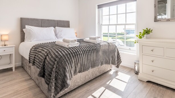 A modern bedroom with a large bed, white nightstands, a dresser, and a window with a view of greenery. - 4-Bedroom Seafront House in Southend | Sea Views - Pass the Keys (Southend-on-Sea)