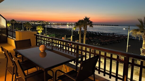 A balcony with a table and chairs overlooking a beach and ocean at sunset. - Beachfront Paradise (Rethymno)