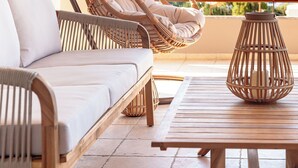 A wicker hanging chair with cushions, a wooden sofa, and a wooden table on a balcony with a view of a hilly landscape.