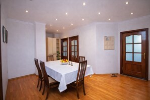 A dining room with a wooden table, chairs, and a fruit bowl. There is a wooden door and a bulletin board on the wall.