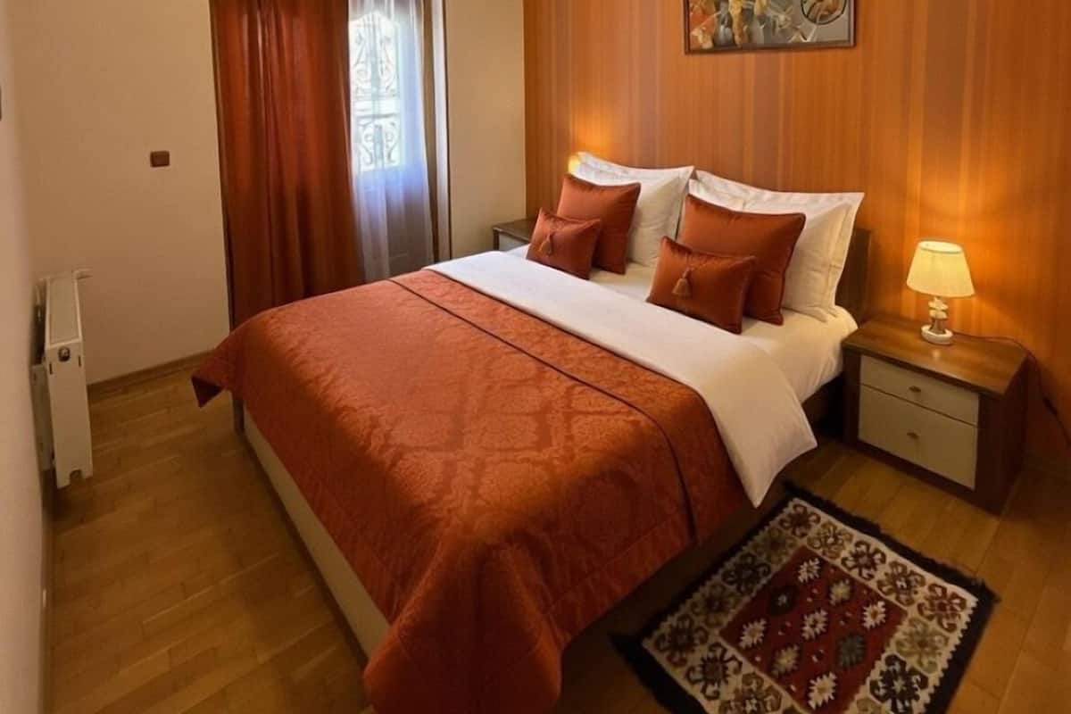 Ein Schlafzimmer mit Bett, Nachttisch, Lampe, einem Fenster mit Vorhängen, einer Holzwandverkleidung und einer Deckenleuchte.