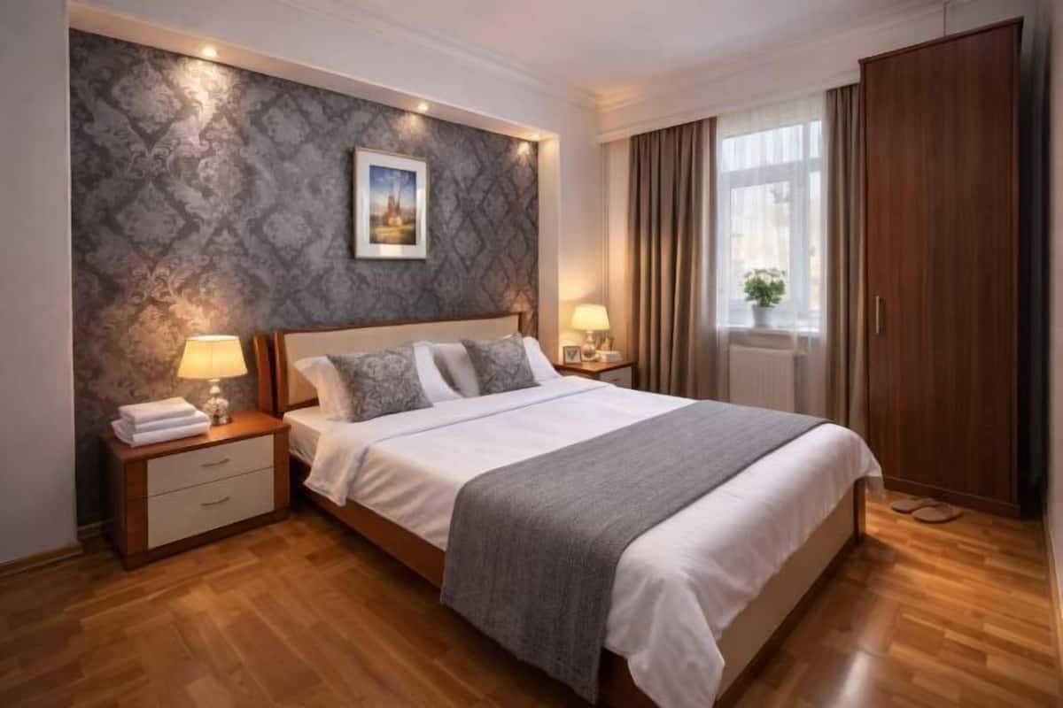 Ein Schlafzimmer mit einem großen Bett, zwei Nachttischen mit Lampen, einem Kleiderschrank, einem Fenster mit Vorhängen und einer Wand mit Muster.