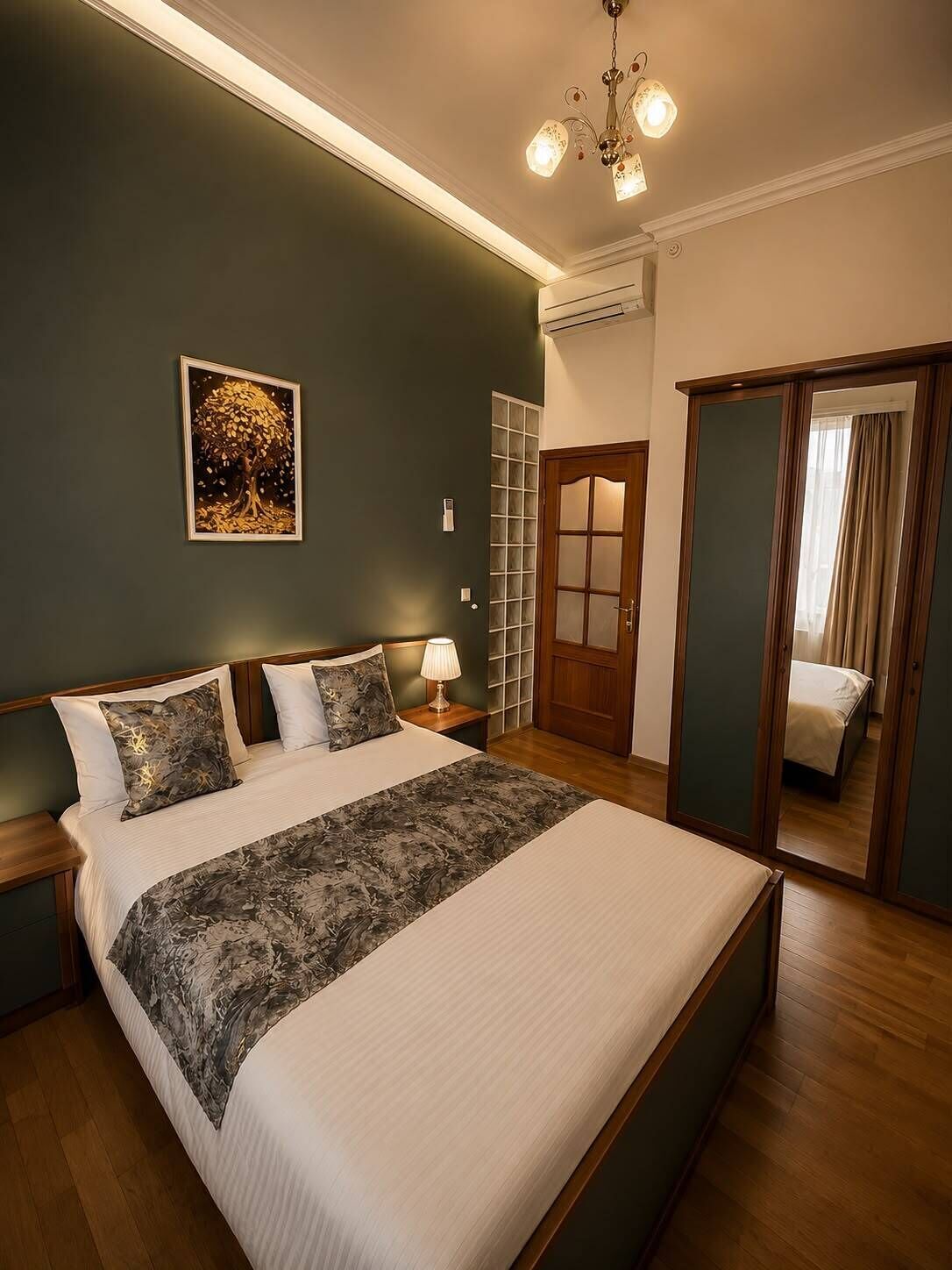 A bedroom with a large bed, a nightstand, a wall-mounted air conditioning unit, a framed artwork, and a sliding door leading to another room.