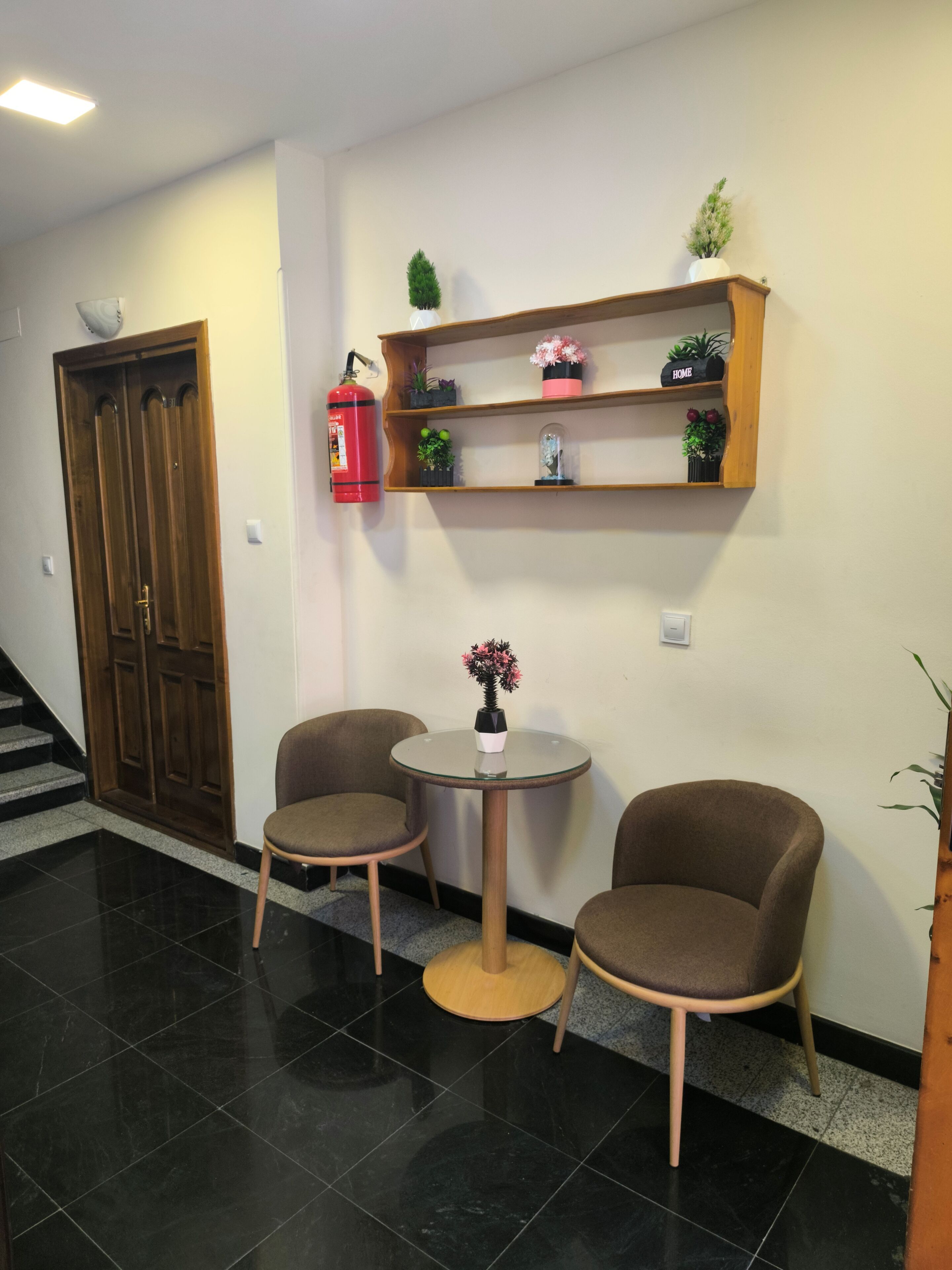 Un espace d’accueil doté d’une porte en bois, d’un extincteur, d’une petite table avec des chaises et d’une étagère ornée d’objets décoratifs.