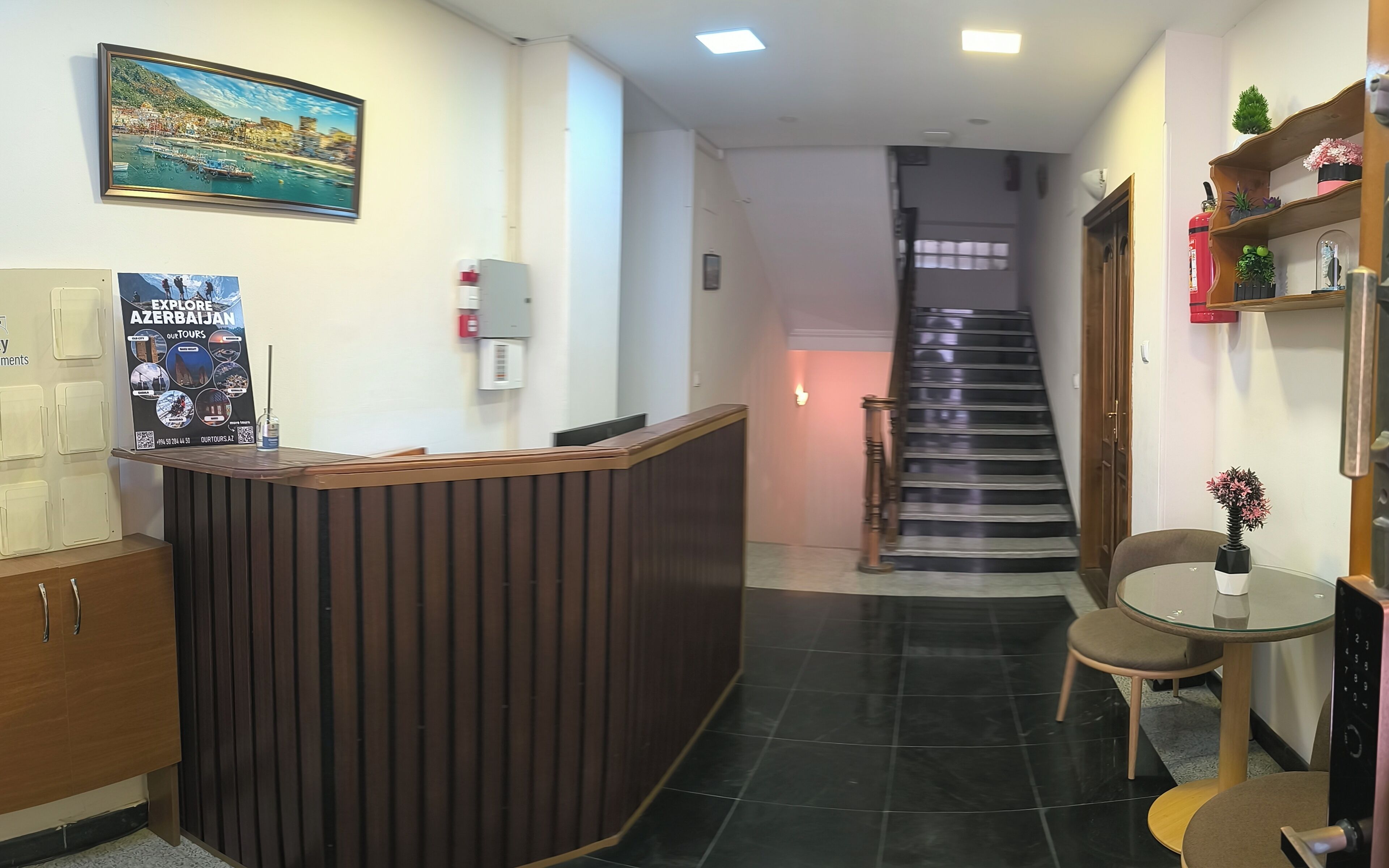 Un espace d’accueil avec un comptoir en bois, une affiche faisant la promotion de voyages en Azerbaïdjan, un escalier et une petite table surmontée d’un vase.