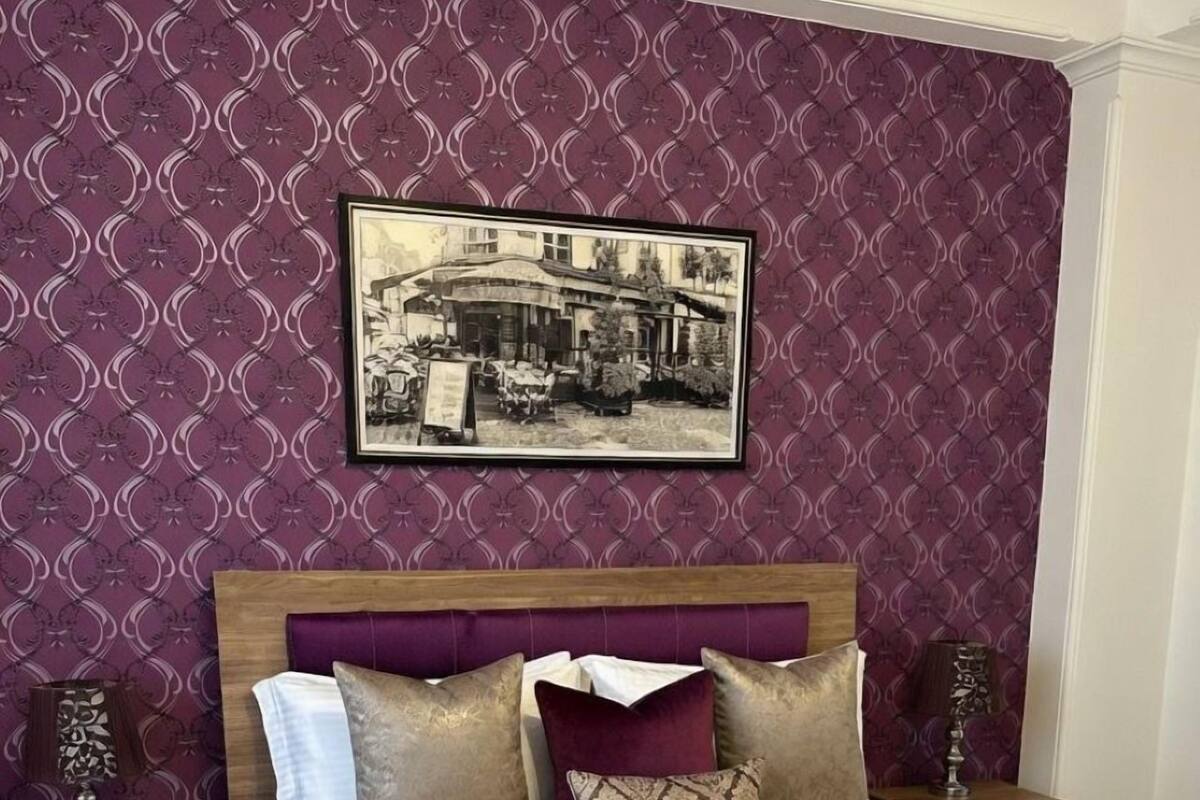 Ein Schlafzimmer mit einem gemusterten Wandbehang, einem Bett mit vielen Kissen, einem Kopfteil, einem Nachttisch mit Lampe und einem gerahmten Bild an der Wand.