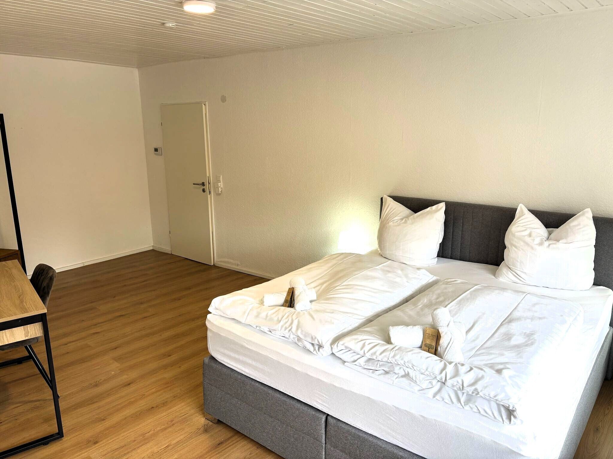 A neatly made bed with white linens and pillows, a wooden floor, a grey upholstered headboard, and a small table with a chair.