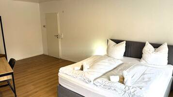 A neatly made bed with white linens and pillows, a wooden floor, a grey upholstered headboard, and a small table with a chair.