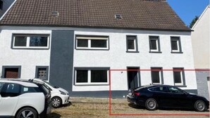 Ein zweistöckiges Haus mit Ziegeldach und Schornstein. Vor dem Haus befinden sich zwei parkende Autos.