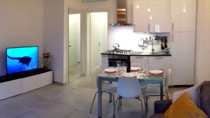 Un salon moderne avec un téléviseur, une table à manger pour deux personnes et un canapé agrémenté d’un coussin jaune.