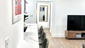 Ein modernes Wohnzimmer mit Fernseher, ein Essbereich mit weißem Tisch und grauen Stühlen und ein Flur, der in einen anderen Raum führt.