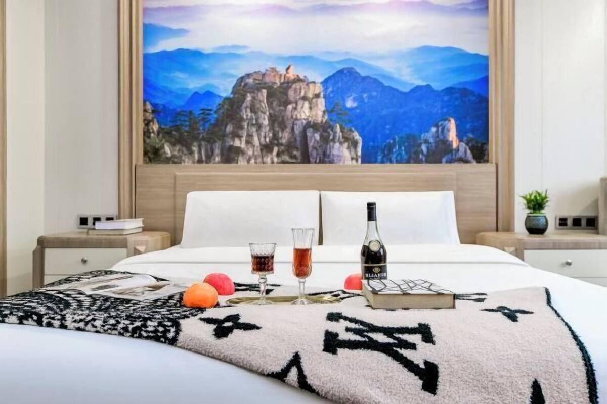Een net opgemaakt bed met een berglandschap aan de muur, een fles wijn, twee glazen en een boek op het bed.