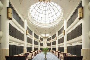 Un vestíbulo de hotel con una lámpara de araña central, techos altos y una disposición de asientos simétrica.
