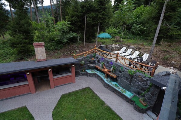A patio area with a stone waterfall feature, lounge chairs, and a wooden deck. - Hotel EverGreen (Borjomi)