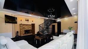 A modern living room with a fireplace, white leather sofas, a black spiral staircase, and a flat-screen TV mounted on the wall. - Hotel EverGreen (Borjomi)