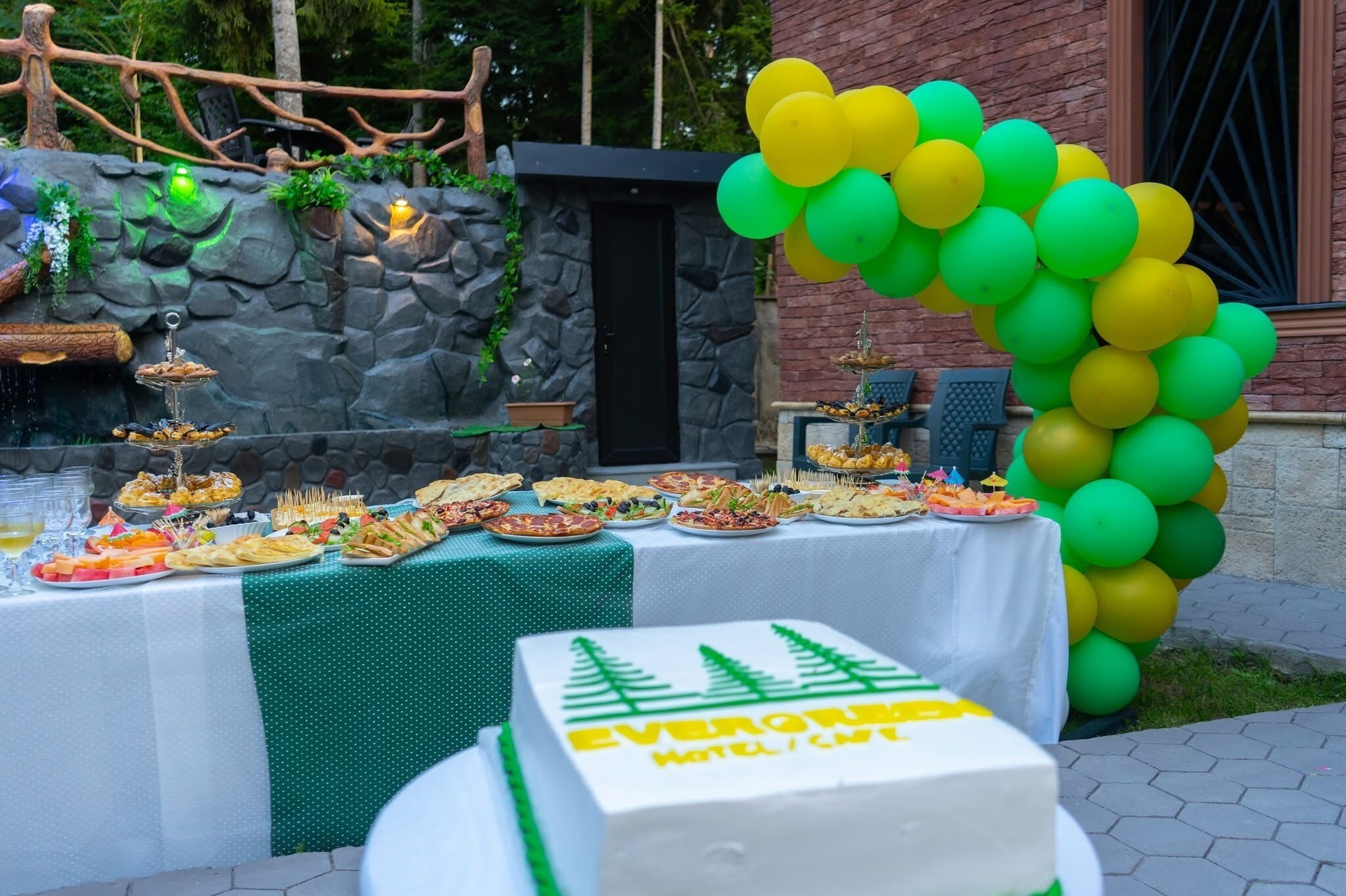 Una mesa de buffet con diferentes comidas, un arco decorativo de globos y un pastel con la inscripción 'EVERGREEN HOTEL'.