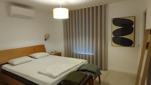 Habitación de hotel con cama, mesita de noche, lámpara, aire acondicionado, cortinas y un cuadro en la pared.