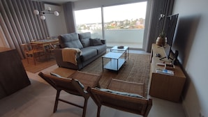 Una sala de estar moderna con un sofá, una mesa de centro y sillas. Hay un comedor con una mesa y sillas. Un televisor está montado en la pared, y una ventana ofrece vistas a la ciudad.