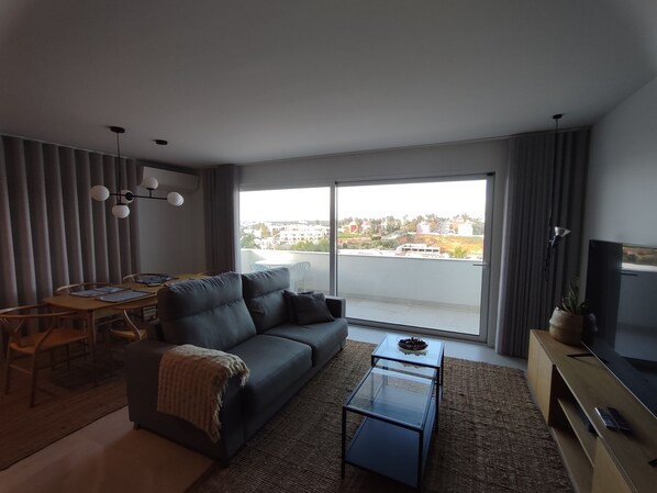 A modern living room with a sofa, a glass coffee table, a TV, and a balcony with a city view. - Terrace Ferragudo River View Flat Super (Lagoa)