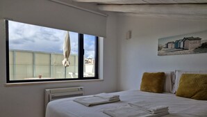 Un dormitorio con un ventanal grande, un radiador, una cama bien hecha con toallas, y un cuadro de cabañas en la playa en la pared.