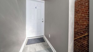 A narrow hallway with a white door marked with the number 4, a doormat, and a wooden handrail against a brick wall. - Queen City Views Suites (Cincinnati)