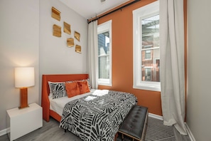 A bedroom with a bed, a nightstand, a bench, and a window with curtains. - OTR 1BR Private Balcony Near Downtown (Cincinnati)