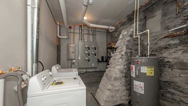 A utility room with a large water heater, a washing machine, and various pipes and electrical panels.
