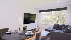 Ein modernes Wohnzimmer mit Essbereich, einer Couch, einem an der Wand befestigten Fernseher und einem Fenster mit Blick auf Bäume.