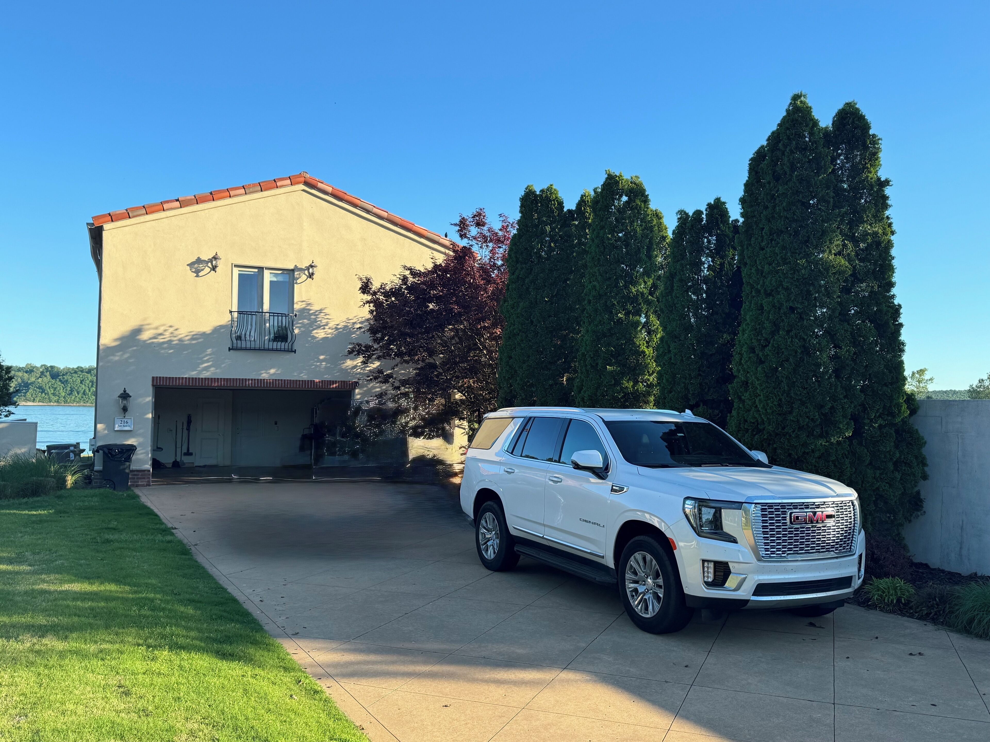 A white GMC SUV parked in front of a two-story house with a garage, a small porch, and a view of a lake.