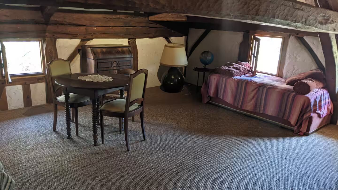 A rustic attic room with a wooden table, two chairs, a lamp, a globe, and a bed with a striped blanket.