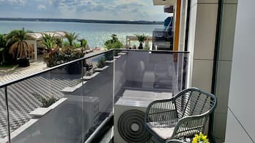 A balcony with a glass railing overlooking a cityscape and a body of water. There are two wicker chairs and a small table with a cushion.