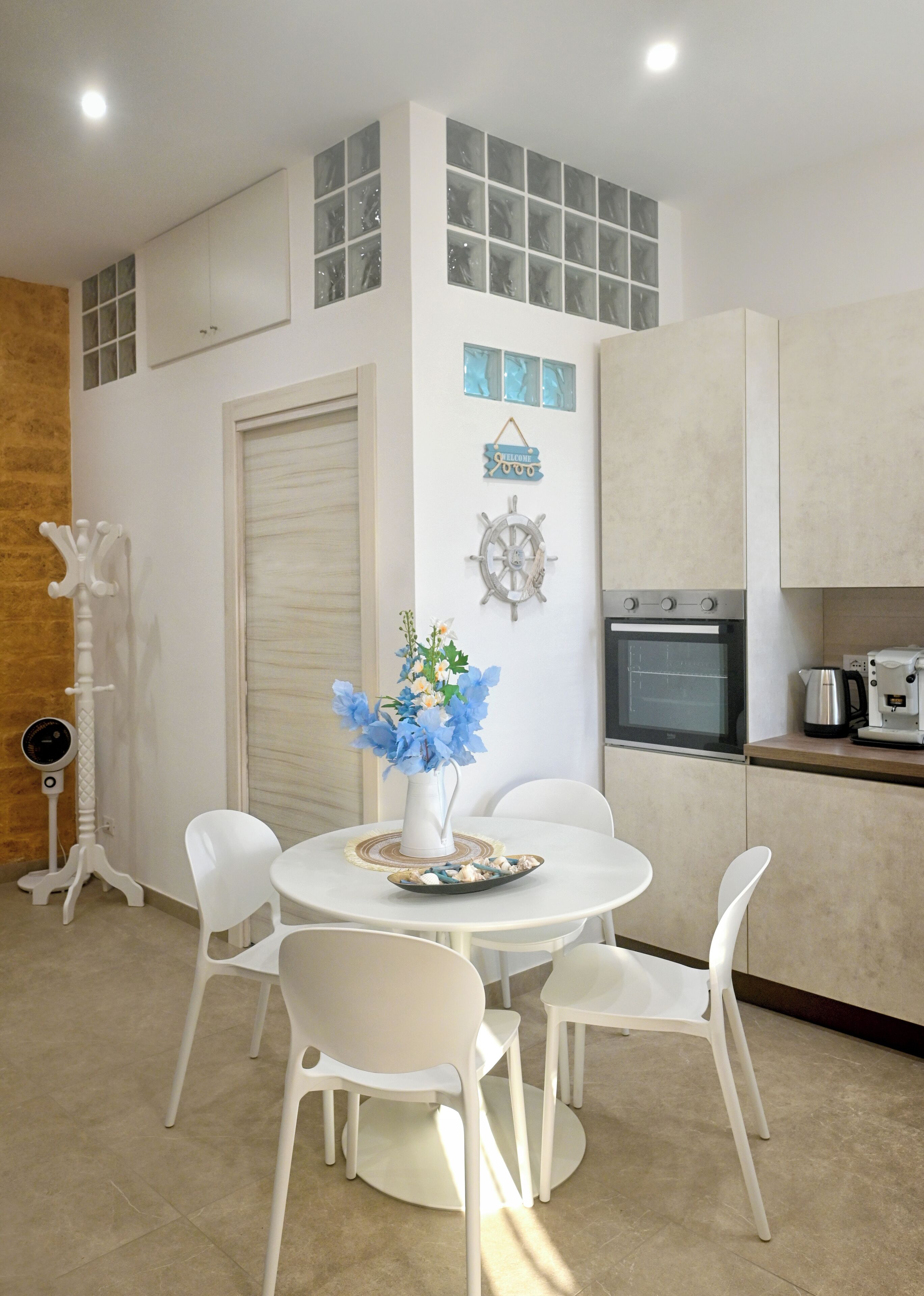 A dining area with a round table, white chairs, a vase with flowers, and a ship wheel decoration.