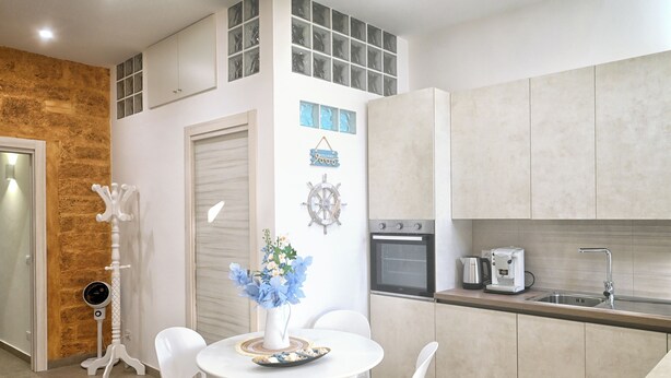 A modern kitchen with white cabinets, a built-in oven, and a coffee maker. There is a dining area with a white table and chairs, and a vase with flowers on the table.