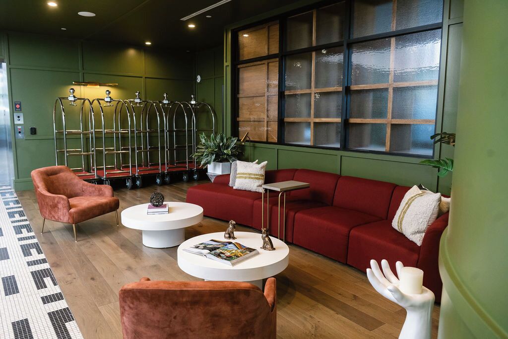 A modern lounge area with a red sofa, two round coffee tables, and a pink armchair. There are metal chairs in the background and a large window with a view.