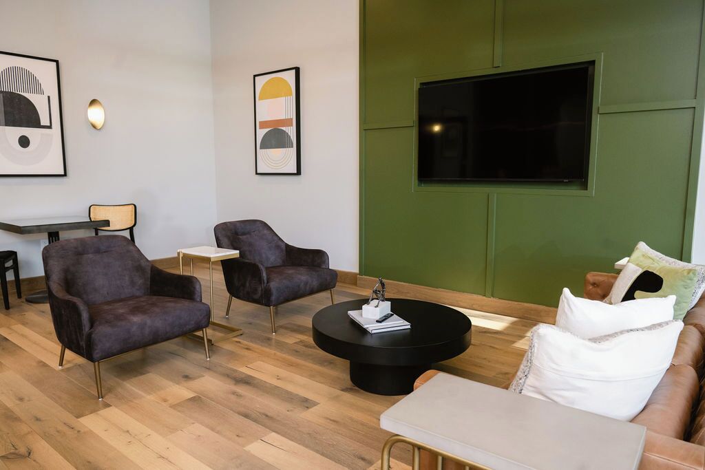 A modern lounge area with two armchairs, a round coffee table, a sofa, and a TV mounted on a green wall.