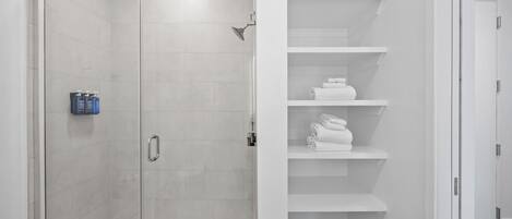 A modern bathroom with a glass-enclosed shower, white tiled walls, and a built-in shelf with folded towels.