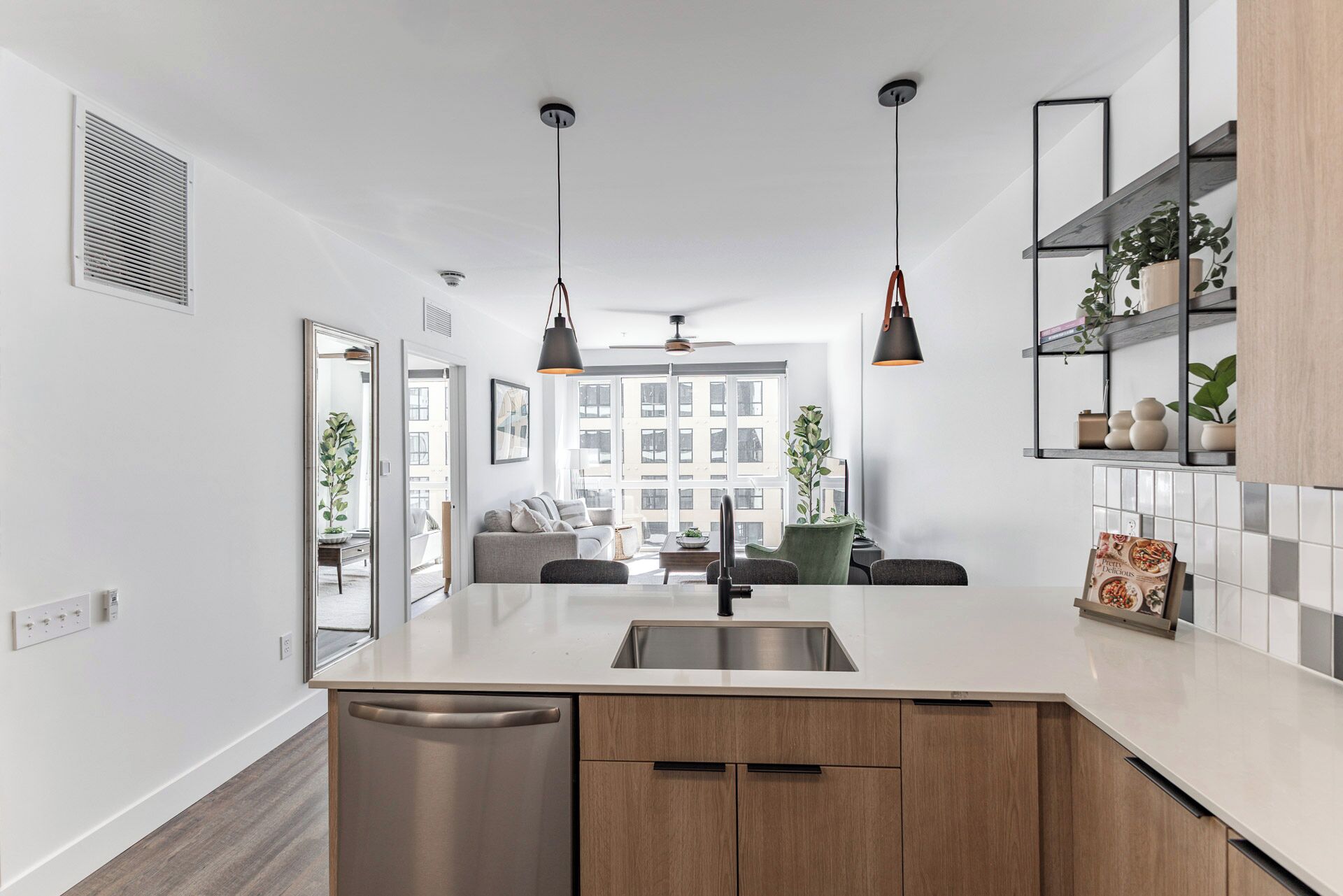 A modern kitchen with a central island, stainless steel sink, and wooden cabinets. There are pendant lights, a ceiling fan, and a view of the city through large windows.